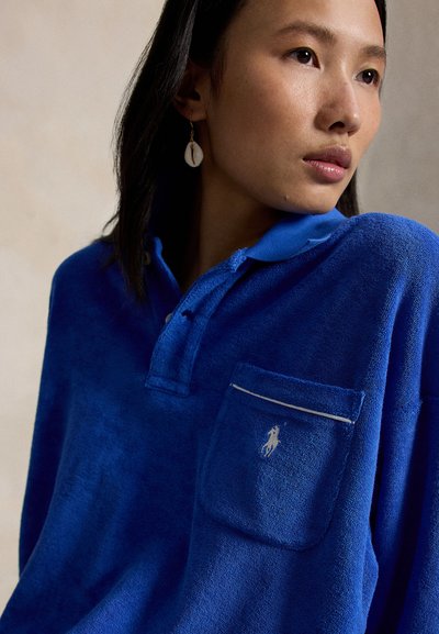 Mujer con polo azul texturizado con un pequeño logotipo bordado en el bolsillo del pecho y un pendiente en forma de concha, mirando hacia la derecha.