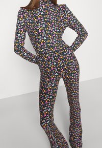 Eine Frau trägt einen taillierten, langärmeligen Blumen-Jumpsuit mit hochgeschlossenem Kragen, der kleine pinke, gelbe, weiße und lila Blumen auf schwarzem Hintergrund zeigt.