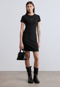 Μαύρο φόρεμα με κοντά μανίκια και στενή γραμμή, διακριτικό λογότυπο στο στήθος, συνδυασμένο με μικρό μαύρο τσαντάκι και μαύρες μπότες μέχρι το γόνατο.