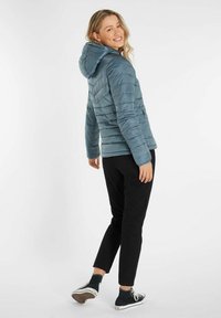 Hellblaues Daunenjacke mit Kapuze, mit horizontalem Steppdesign, mit Reißverschlusstaschen, getragen über schwarzen Hosen und Schuhen.