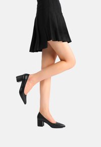 Pump in pelle nera con tacco alto, punta appuntita e tacco quadrato, caratterizzate da una texture liscia e da una silhouette elegante.