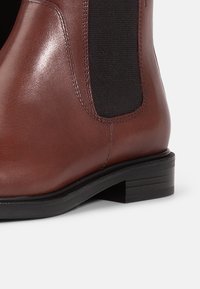 Bottine en cuir marron avec panneaux latéraux élastiques, petit talon empilé et finition lisse. Elle présente un accent textile noir contrastant.