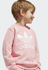 Rúžová mikina vyrobená z jemného materiálu, s veľkým bielym logom Adidas a dizajnom troch pruhov na prednej strane. Okrúhly výstrih a dlhé rukávy.