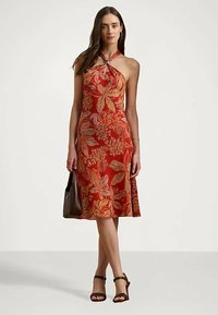 Moteris vilkinti raudoną suknelę su apykakle ir oranžiniais gėlių raštais, laiko rudą rankinę ir vilki juodas kulkšnines žemakulnes.