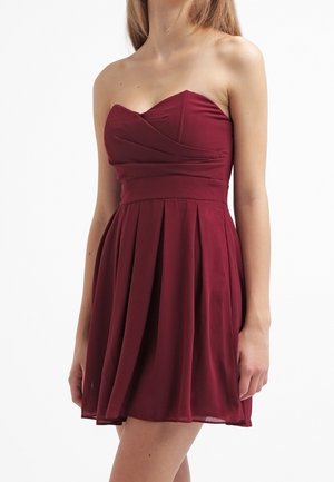 Vrouw draagt een strapless, bordeaux Rode geplooide mini-jurk, staand tegen een witte achtergrond, zichtbaar van nek tot midden bovenbeen.