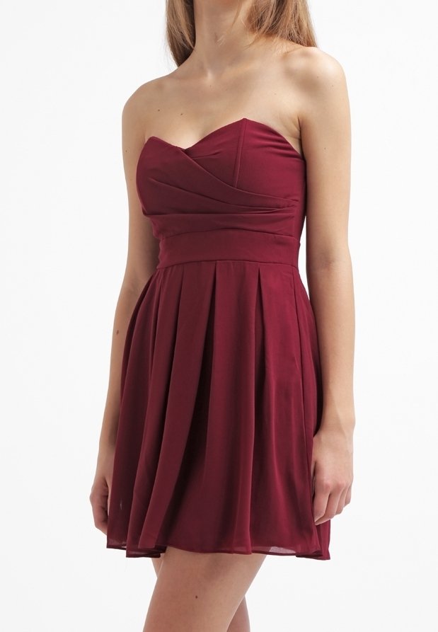 Femme portant une mini-robe plissée bordeaux sans bretelles, debout devant un fond blanc, visible du cou jusqu'à mi-cuisse.