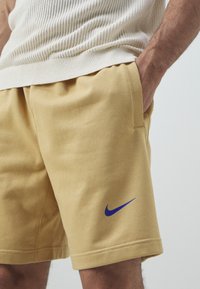 Mężczyzna ubrany w beżowe szorty Nike z niebieskim logo swoosh, ręka w kieszeni, a na górze widoczny kremowy sweter od talii do połowy uda.