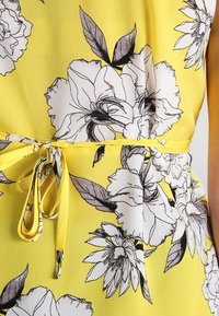 Tessuto giallo con grandi motivi floreali neri e bianchi, con una sottile cintura annodata in vita su un capo senza maniche.