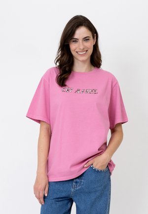 Jonge vrouw lacht, draagt een roze t-shirt met de tekst "OH APRIL" en een blauwe spijkerbroek, staat met één hand in de zak tegen een witte achtergrond.