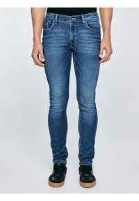 Vīrietis, kurš valkā šauras zilās džinsa bikses un melnus sporta apavus ar brūnām zolēm, stāv pret vienveidīgu gaišu fonu.