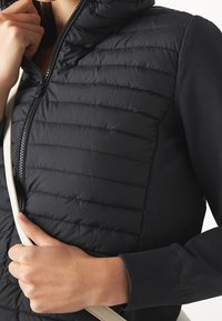 Persona que lleva un chaleco negro acolchado con cremallera, sosteniendo una correa de bolsa beige y ajustando el cuello del chaleco con una mano.