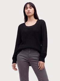 Junge Frau steht, trägt einen schwarzen Waffelstrick-Langarmpullover und graue Hose vor einem schlichten hellen Hintergrund.