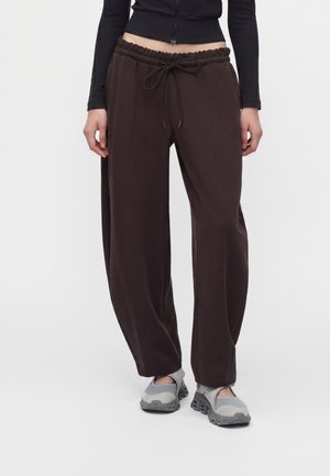 Persoană purtând pantaloni sport largi maro închis cu șnur în talie, bluză neagră scurtă cu mâneci lungi și pantofi gri slip-on, stând pe fundal alb.