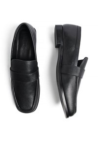 Melnas ādas mokasīni ar kvadrātu formas purngaliem, ar platu siksnu pāri augšdaļai un zemu bloku papēdi. Gluda apdare un minimālisma dizains.