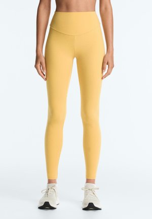 Žena v žltých legínach s vysokým pásom a bielych teniskách stojaca pred jednoduchým svetlým pozadím.