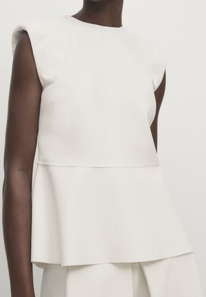 Dark-skinned person wearing sleeveless white layered top and matching white pants against plain light background.