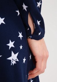 Hand som vilar mot marinblått tyg med vita stjärnmönster; ärmkaftet har en liten knapp och en slitsöppning.