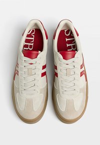 Baskets en daim beige et rouge avec un design à lacets, comportant des accents rouges et une semelle en caoutchouc naturel. L'intérieur présente un logo imprimé en rouge.