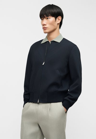 Μπλε ζακέτα με φερμουάρ, με ανοιχτοπράσινο γιακά, δύο πλαϊνές τσέπες και ριμπ μανσέτες· κατασκευασμένη από λείο ύφασμα με μινιμαλιστικό σχέδιο.