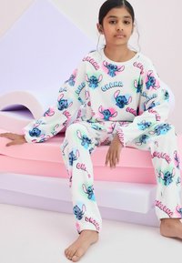 Valkoiset fleece-yöpuku, joissa on suuria, värikkäitä Stitch-hahmotulosteita ja "Ohana"-teksti. Löysä malli elastisilla hihoilla, pastellitaustalla.