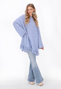 Világoskék, túlméretezett kötött pulóver bojtós szegéllyel, amelyet világos farmerudvarú, bő szárú nadrággal párosítanak. Simább textúra és laza fazon.