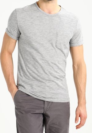 Homme portant un t-shirt gris clair à manches courtes avec une poche avant et un pantalon gris foncé, debout avec une main dans la poche.