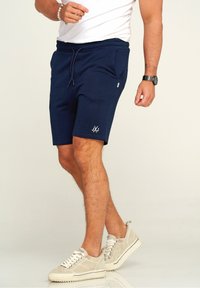 Navy blue shorts in a soft fabric, featuring an elastic waistband with drawstring, side pockets, and a white logo on the lower leg.
