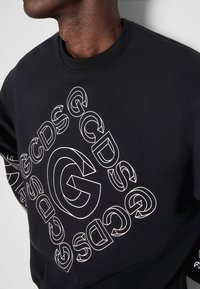 Personne portant un sweat-shirt noir avec "GCDS" en contour blanc répété en motif en spirale sur le devant et la manche.