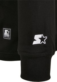 Manche de sweat-shirt noire avec bord-côte présentant le logo étoile blanc de la marque Starter et un patch rectangulaire Starter Black Label.