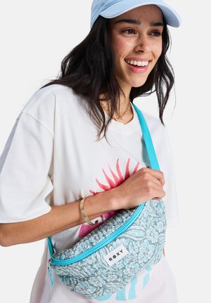 Lächelnde Frau trägt eine hellblaue Mütze und ein weißes T-Shirt, hält eine blaue Roxy-Umhängetasche mit Blumenmuster und trägt ein goldenes Armband am Handgelenk.