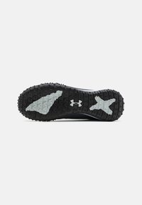 Suela de zapato atlético negro con textura de goma prominente, que presenta un distintivo logo de Under Armour y acentos en gris claro.