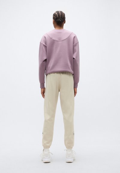 Μωβ φούτερ με καμπυλωτή ραφή στο λαιμό, συνδυασμένο με μπεζ στενά παντελόνια φόρμας. Άσπρα αθλητικά παπούτσια ολοκληρώνουν την εμφάνιση.