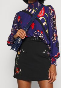 Blusa morada con estampado floral y cuello alto, combinada con una falda negra con bordados de colores y una cinturilla con tachuelas; sosteniendo un bolso azul.