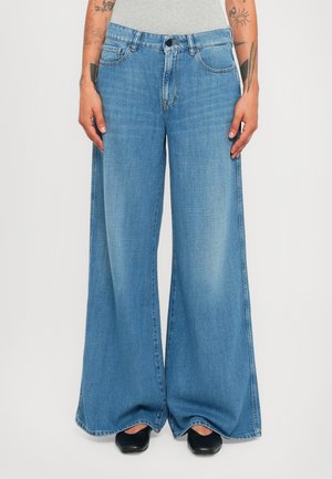 Personne portant un jean large bleu clair, un haut blanc et des chaussures noires, se tenant les mains détendues sur les côtés sur un fond uni.