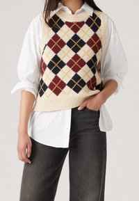 Débardeur en maille avec un motif en losange crème, marine, bordeaux et jaune. Bords côtelés, porté sur une chemise blanche à col et un jean foncé.