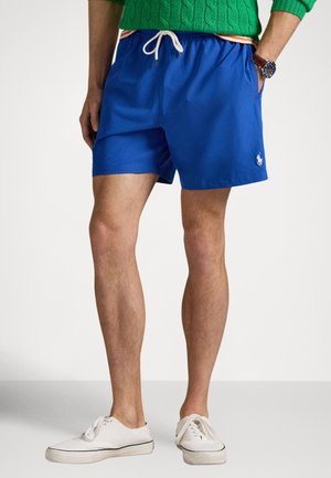 Modré plavkové šortky s elastickým pásom a bielym sťahovacím šnúrkou, so bočnými vreckami a malým logom na leme. Nosia sa s bielymi topánkami.