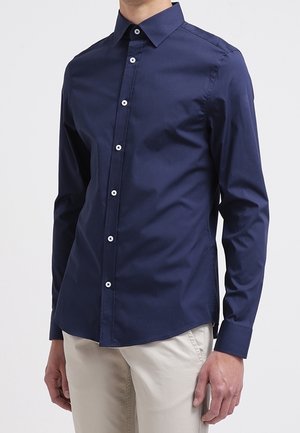 Mann trägt ein eng anliegendes, navyblaues Hemd mit langen Ärmeln und Knöpfen sowie hellbeige Hose, steht mit entspannten Armen seitlich am Körper.