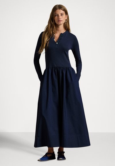 Μακρύ σκούρο μπλε φόρεμα με μακριά μανίκια, κουμπωτή πατιλέτα και διακόσμηση με βολάν στον λαιμό. Σούρες στη μέση και δύο πλαϊνές τσέπες. Φορεμένο με μπλε παπούτσια.