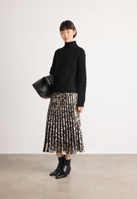 Vrouw in een zwarte turtleneck trui, geplooide rok met luipaardprint, zwarte enkelboots, houdt een zwarte leren tas vast tegen een vlakke muur.