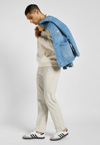 Felpa con cappuccio tan, pantaloni in corduroy beige e sneakers bianche con strisce nere. Giacca di denim appoggiata sulla spalla, con una tasca.