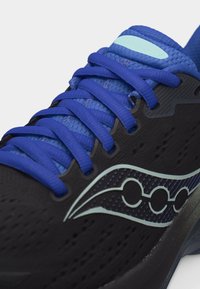 Αθλητικό παπούτσι με μαύρο επάνω μέρος, μπλε κορδόνια, μπλε επενδυμένη γλώσσα και λογότυπο ανοιχτού μπλε στο πλάι, εμφανίζεται σε κοντινή γωνιακή όψη.