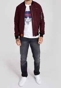 Giubbotto bomber color bordeaux con zip frontale e polsini elasticizzati. Sotto una maglietta grafica bianca, abbinata a jeans grigi e sneakers bianche.