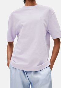 Lavendelfärgad bomull t-shirt med rund halsringning, kort ärm och en subtil broderad logotyp på bröstet, ihop med ljusblå byxor.