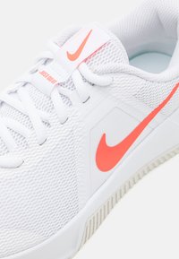 Hvide atletiske sko med en struktureret mesh-overdel, kontrastende orange Nike swoosh og lys sål. Snørebånd og rem er til stede.