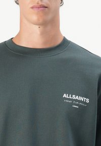 Sweatshirt verde escuro com decote redondo e textura canelada. Apresenta a marcação em texto branco "ALLSAINTS" e caracteres japoneses no peito.