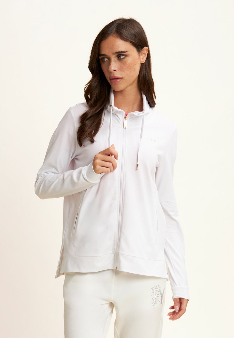 Giacca sportiva bianca con zip, colletto alto, cordino e dettagli in oro. Presenta una texture liscia e un design aderente.