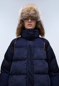 Námořnická bunda s prošívanými horizontálními panely, vysokým límcem a kapucí lemovanou kožešinou. Texturované tkaniny s koženými detaily.