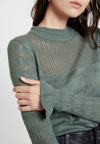 Zelený pletený svetr s texturovaným vzorem a dlouhými rukávy s rozšířenými manžetami, který se pyšní otevřenými krajkovými detaily na ramenou a trupu.