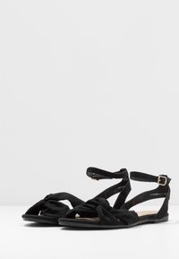 Sandales en daim noir avec un design à l'avant noué, des brides fines autour de la cheville et une semelle plate. Comprend une petite boucle dorée pour l'ajustement.