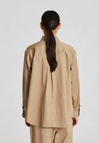Beige, ylisuuria paita pehmeästä kankaasta, pitkät hihat, napin yksityiskohdat hihansuissa ja pyöristetty helma, näkyy takaa.
